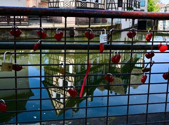 Ein Metallzaun mit absplitternder roter Farbe, der mit zahlreichen roten herzförmigen Vorhängeschlössern bestückt ist. In einem ruhigen Fluss spiegelt sich der Hintergrund der Szenerie, hübsche Fachwerkhäuser.