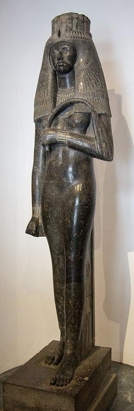 a statue, it is a slender woman with a large queen's headdress, she is holding a horse whip, her one hand at her abdomen, the other at her side