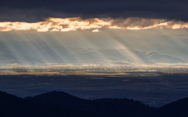 Blick vom Schwarzwald über die Rheinebene zu den Vogesen, mit Teleobjektiv aufgenommen. Den Vordergrund bilden dunkle Schwarzwaldhügel, die im Schatten liegen. Dahinter liegt die Rheinebene, die in zwei kontrastierende Hälften geteilt ist: vorne im Schatten, hinten sonnendurchflutet. Darüber sind die Hügel der Vogesen im Dunst zu erkennen. Am oberen Bildrand hängt eine dunkle Wolkendecke, deren unterer Rand von hinten durch die Sonne angestrahlt ist. Das Sonnenlicht, das die Landschaft darunter beleuchtet, bildet ausgeprägte Strahlen im Dunst.
