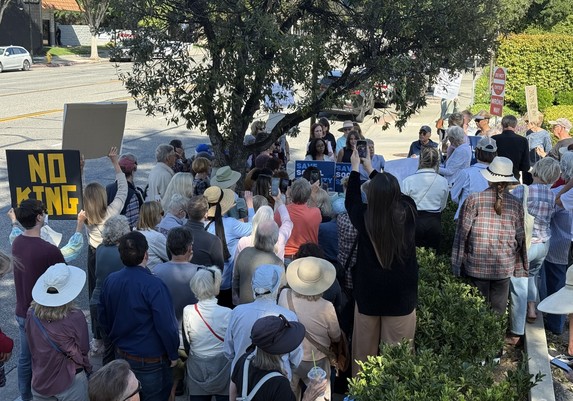 A large group of people gathered at a protest in Thousand Oaks, holding signs with messages such as "NO KING" and "SAVE SOCIAL SECURITY." They are assembled around speakers, with many attendees recording the event on their phones.