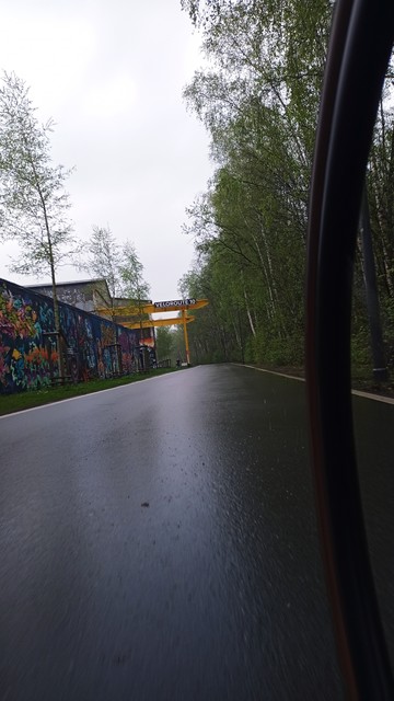 Ein Vorderradreifen eines Fahrrads in voller Fahrt auf einer breiten Veloroute. Voraus ein gelbes Metallkonstrukt mit der Aufschrift "Veloroute 10", links am Fahrbahnrand viele Gebäuderückseiten mit farbenfrohen Graffitti. Es ist bewölkt und naß.