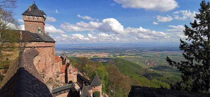 Vue du haut d'un château fort en pierres rougeâtres en premier plan sur la gauche, avec en arrière plan une plaine avec des champs colorées, en arrière plan très loin on devine des collines. Le ciel est bleu avec des nuages.