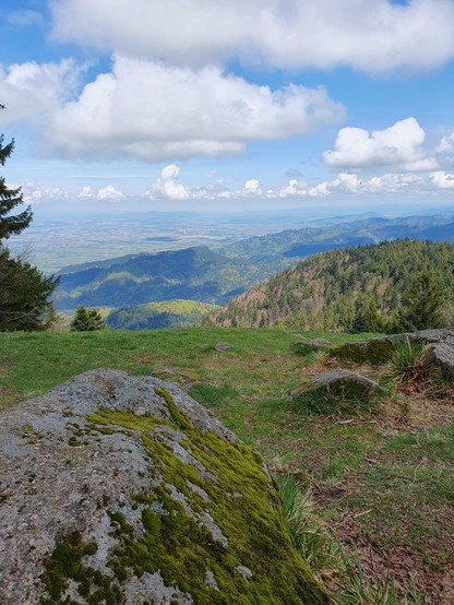 im Vordergrund ein bemooster Felsen, dann ein abfallender Hang mit magerem Rasen, dahinter sieht man den Blick vom Berg hinunter auf weitere bewaldete Berge, am Himmel einige Wolken, die ihre Schatten auf den Wald darunter werfen