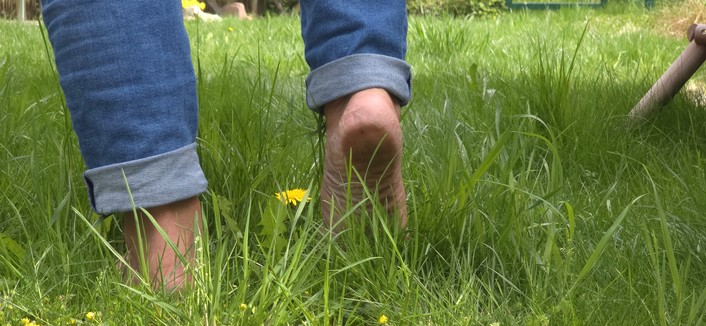 Ein Mann, von dem nur der untere Teil der Waden und Füße zu sehen ist, läuft barfuß durch grünes Gras vom  Betrachter weg. Der rechte Fuß hebt vom Boden ab und die Fußsohle an drr Ferse ist zu erkennen, während der Fußballen noch im dem Gras steht.