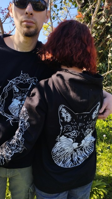 Sur la photo une femme est de dos et porte un sweat shirt noir avec une tête de renard imprimée en blanc. Sur son bras gauche on voit une branche de lierre imprimée en blanc.À côté d'elle un homme de face porte un tee shirt noir avec une tête de renard blanc imprimé dessus, entourée d'entrelacs végétaux et de chêne.