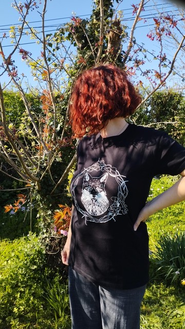 Photo d'une femme se tenant près d'un arbre dans un jardin, elle porte un tee shirt noir avec une tête de renard et des entrelacs végétaux imprimés en blanc. Tee shirt unisexe.