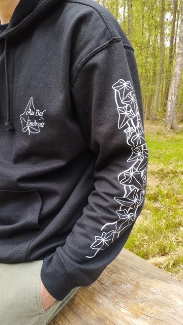 Détail des manches et de l'avant du sweat shirt noir. On y voit la branche de lierre imprimée en blanc sur le bras gauche d'un homme assis sur un tronc d'arbre mort. Sur l'avant cœur du sweat, est imprimé en blanc le logo au bel endroit entouré d'une feuille de lierre