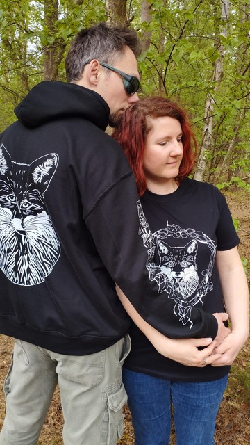 Photo d'un couple en forêt portant pour lui le sweat shirt noir avec la tête de renard en blanc dans le dos et pour elle le tee shirt noir avec la tête de renard et entrelacs de végétaux imprimés en blanc sur le devant du tee shirt