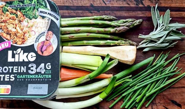 auf einem Brett eine Packung LIKE PROTEIN BITES Gartenkräuter, einige Stangen grüner Spargel, Frühlingszwiebeln, 1 Karotte 1 Pastinake 1 Stück Porree, einige Zweige Salbei und Schnittlauch