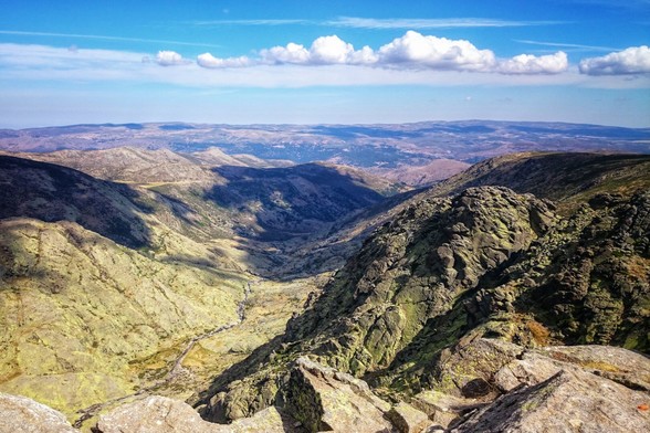 A wide shot from a high vantage point overlooking a vast mountain range with valleys and a partly cloudy sky.