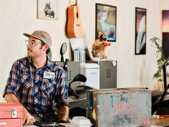 Man in room with artworks and a guitar hanging on the walls. He's looking to one side and sat in front of a table/desk that has a sign kit box on it that is lettered with "My signs don't cost, they pay".