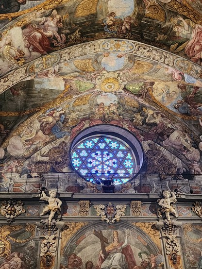 Wall and arched/vaulted ceiling of a church, covered with frescoes similar in style and subject matter to the Sistine Chapel. Near the top of the arched wall is a round stained-glass window with a six-sided star in mostly blue tones.