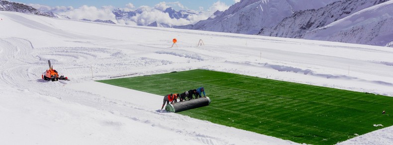 The image shows people rolling out large sheets of green fabric or material over a snowy area. There is snow surrounding the green section, with a snow vehicle nearby. The scene appears to be in a mountainous, snowy landscape, possibly with the material being used to cover or protect part of the snow.