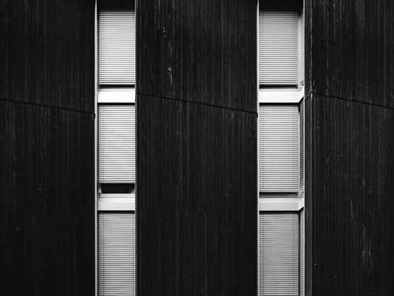 Black and white photo of a modern building facade featuring tall, narrow windows with closed blinds, set against dark vertical wooden panels. The strong lines and repetitive pattern create a minimalist, geometric architectural look.