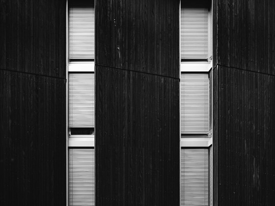 Black and white photo of a modern building facade featuring tall, narrow windows with closed blinds, set against dark vertical wooden panels. The strong lines and repetitive pattern create a minimalist, geometric architectural look.