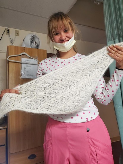 Marietta with a big smile, a young woman with light brown hair in pink and white scrubs and face mask holding up a white lace scarf