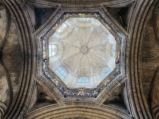 View up into the loggia of the cathedral spire, which is octagonal and has a vaulted ceiling and eight clear glass windows. The loggia is framed by symmetrical ribbed arches on the lower ceiling.