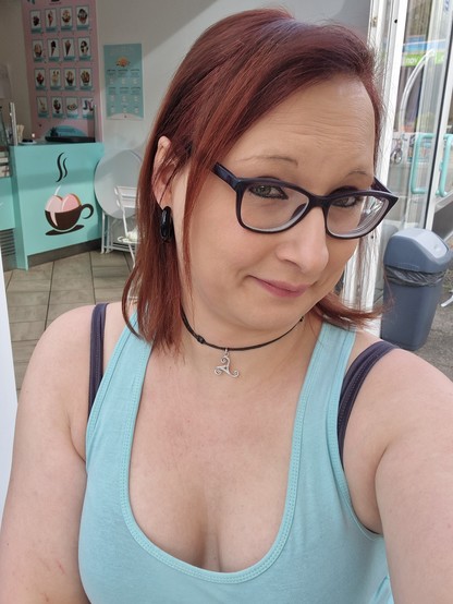 Das Bild zeigt eine Frau mit roten Haaren, die eine Brille mit schwarzen Rahmen trägt. Sie lächelt leicht und trägt eine schwarze Halskette mit einem silbernen Anhänger. Sie trägt ein hellblaues Tanktop mit einem tiefen Ausschnitt. Im Hintergrund ist ein Café zu sehen, das durch eine Glasfront sichtbar ist. Auf der linken Seite des Bildes ist ein Tressen mit einer Illustration zu sehen, und auf der rechten Seite steht eine graue Mülltonne. Die Frau hat einen schwarzen Ohrring im Ohr und wirkt entspannt und glücklich.