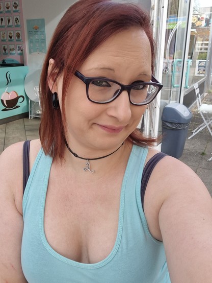 Das Bild zeigt eine Frau mit roten Haaren, die eine Brille trägt. Sie lächelt leicht und blickt direkt in die Kamera. Sie trägt eine schwarze Choker-Kette mit einem silbernen Anhänger, der an eine Spirale erinnert, und einen großen schwarzen Ohrring. Ihre Kleidung besteht aus einem hellblauen Tanktop mit einem tiefen Ausschnitt. Im Hintergrund ist ein Café zu sehen, das durch ein Fenster mit einem Eisbecher-Logo und eine Auswahl an Eiscreme-Sorten gekennzeichnet ist. Die Einrichtung des Cafés ist in einem hellen Blau gehalten, und man kann einen Teil des Innenraums sowie einen grauen Mülleimer im Freien erkennen.