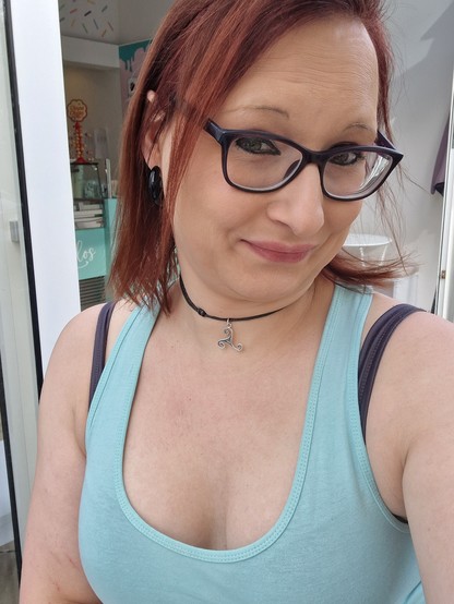 Das Bild zeigt eine Frau mit roten Haaren, die eine Brille mit schwarzen Rahmen trägt. Sie lächelt leicht und blickt direkt in die Kamera. Sie trägt eine schwarze Choker-Kette mit einem silbernen Anhänger, der eine kunstvolle, spiralförmige Form hat. Ihre Ohren sind mit großen, schwarzen Ohrringen geschmückt. Sie trägt ein hellblaues Tanktop mit einem tiefen Ausschnitt, das ihre Schultern und Brust zeigt. Im Hintergrund ist ein Teil eines Gebäudes zu sehen, das möglicherweise ein Geschäft ist, mit einem Tressen, das an eine Eisdiele erinnert. Die Wand ist in einem hellen Blau gehalten und mit bunten Spritzern verziert.