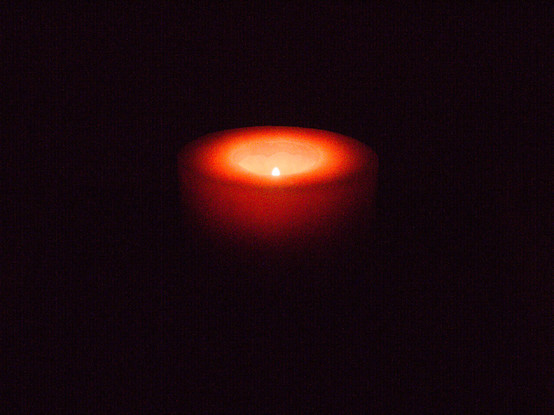 Eine kurze rote Kerze im dunklen Raum mit leuchtender Flamme, die den Kerzenkörper bis zur Höhe des Dochtes rot aufglühen lässt. Wirkt durch Bearbeitung von Schatten und Farbkurve wie weichgezeichnet. Die Flamme ist hell und klar.