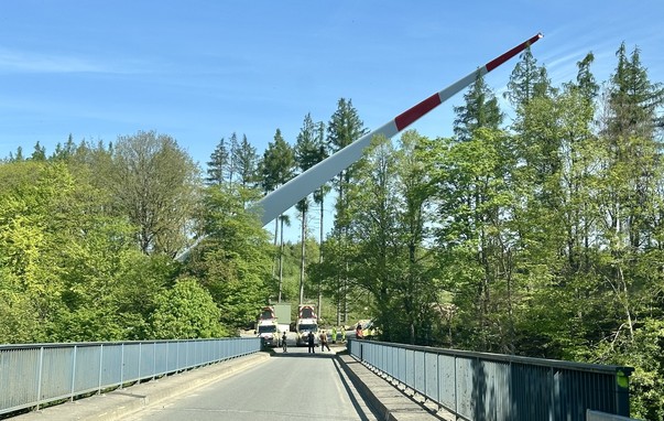 am Ende einer Brücke stehen mehrere Begleitfahrzeuge eines Schwertransports, ein riesiger Windradflügel ragt in die Höhe