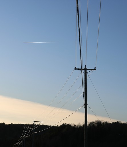 Photo of a small power line taken from under, its five different cables shining slightly in the fading light, and the pole only a dark silhouette against a blue sky. A plane with its white trail is flying in the sky in top left corner. The dark silhouette of the forest is seen in the distance and a white cloud with a triangular shape creates a diagonal line above the horizon.