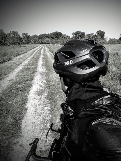 A bike rider seen from behind looking at a gravel road. The road is completly straight. We can see trees in the very background where leads the road. 
Picture is black and white