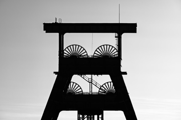 A black-and-white silhouette of a historic mining tower with large pulley wheels, set against a pale sky. The architectural structure is sharply defined, showcasing industrial symmetry and legacy.