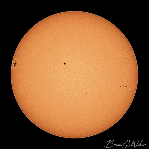 A full disk solar image captured in monochrome and colorized to be orange. Small sunspots dot the surface, but there is a large one coming into view from the left side.