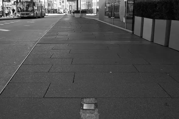 The image shows a city sidewalk with a bus in the background. The pavement has a modern design with rectangular tiles, and there are planters along the edge. It's a black-and-white photo, suggesting a focus on textures and shapes rather than color. The street appears to be relatively quiet, possibly indicating time early in the day or low foot traffic.

Image Credits: Wikimedia / Christian Michelides / CC BY-SA 4.0