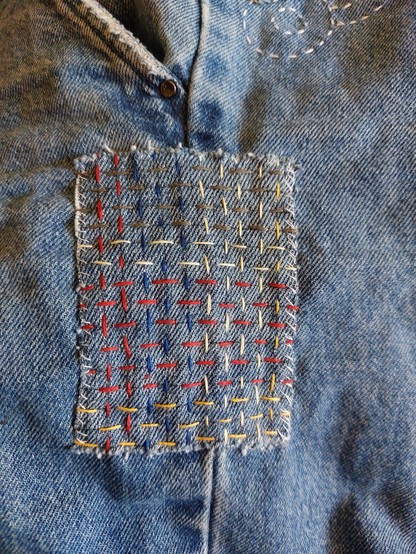 Flicken in Sashiko-Technik mit buntem Stickgarn auf einer Jeansnaht. Die Fäden bilden ein Webmuster.