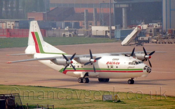Side view of a white, high-winged, 4 propellor-engined cargo plane with a heavily glazed nose cone taxiing from left to right.
A green and red stripe runs along the body, sweeping under the tail, with another, similar stripe running along the base of the tail before sweeping up at the rear.
There are red "Balkan" titles on the upper forward fuselage, under smaller black "Cargo" titles.
Green grass fills the foreground, with grey concrete apron and green grass leading up to a couple of hangars in the distance.
