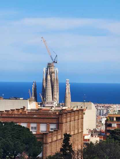 close-up of the the intricate spires of Sagrada familia rising above the cityscape and a building in the foreground. there is a crane on top of the central unfinished spire and the the blue mediterranean sea and hazy sky are in the background.