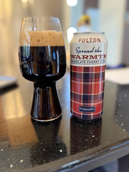 A stout glass of dark beer next to a pint can wrapped in a Faribault Mill plaid red pattern