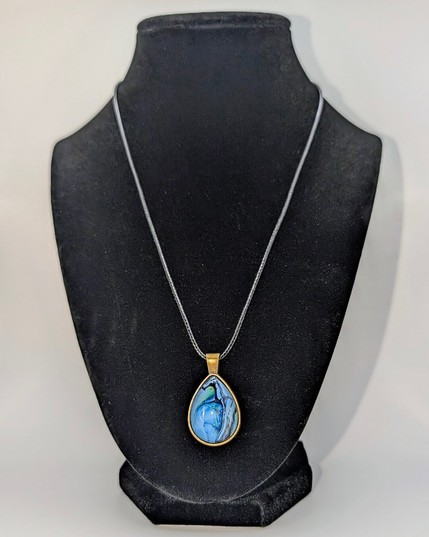 A teardrop-shaped pendant necklace displayed on a black jewelry mannequin. The setting is bronze and the design is primarily light blue.