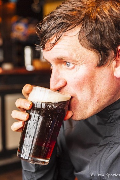 A man with light skin, slightly flushed, and dark hair takes a first sip at a pint of dark beer in a straight glass. He faces left and his face is softly light by light from a window, which can be seen reflected in the glass.
