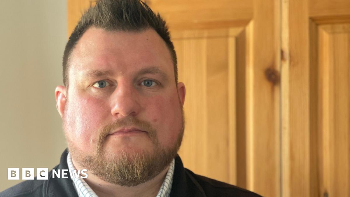 Snagger Chris Greenwood is wearing a checked shirt with a black fleece gilet over the top. The background is a wooden door and pale coloured painted wall.