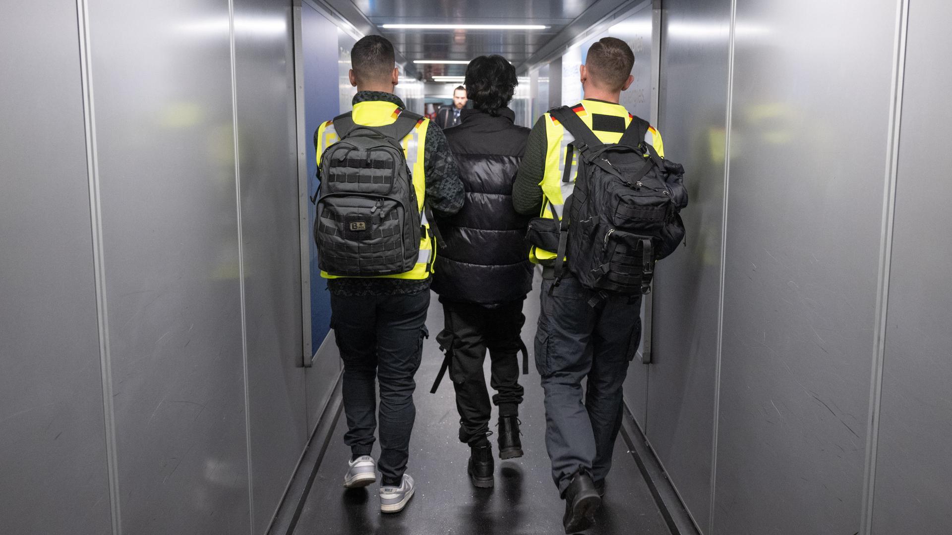 Zwei Beamte in Warnwesten eskortieren einen Mann durch einen Flugzeugtunnel.
