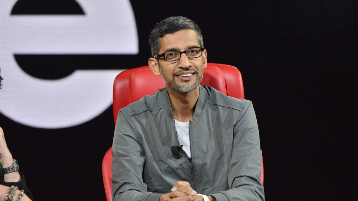Google CEO Sundar Pichai smiling on stage at a conference where he was speaking. He sits in a comfortable chair and is leaning over with his hands clasped together.