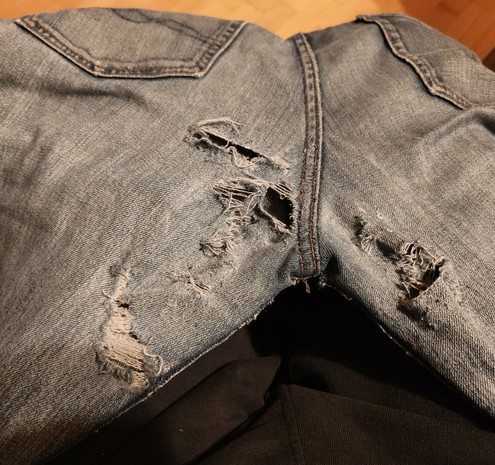 Foto von einer blauen Jeans. Gezeigt wird das Gesäß Unterhalt der Gesäßtaschen. Entlang der Mittelnaht der beiden Beine ist der Stoff an diversen Stellen eingerissen, an anderen stellen bereits fadenscheinig geworden.  