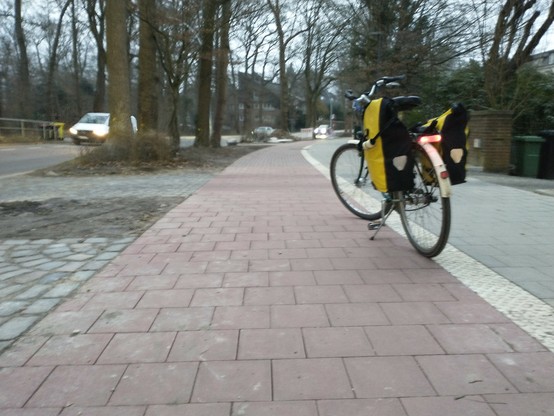 Ein Fahrrad mit gelben Taschen steht am Rand eines rot gepflasterten Radweges, daneben ein grau gepflasterter Gehweg. Rechts und links davon stehen große Bäume, links ist die Fahrbahn zu sehen.