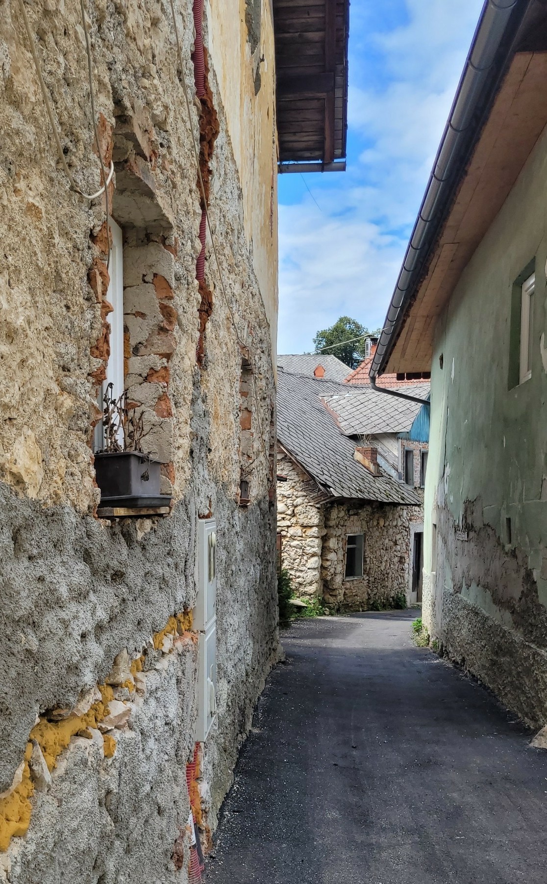 Eine schmale, asphaltierte Straße, die sich nach rechts windet. Links und rechts stehen direkt anschließend alte Häuser. Die Mauer des ersten Hauses auf der linken Seite ist wegen Renovierungsarbeiten nicht verputzt und besteht aus rauhem Stein und einer bauschaumartigen Substanz. Auf der Fensterbank eines kleinen Fensters steht ein Blumenkasten mit einer verdorrten Pflanze. Das zweite Haus dort ist ebenfalls nicht verputzt und man sieht, dass es aus Naturstein gebaut wurde. Es ist mit alten, flachen, anthrazithgrauen Schindeln gedeckt. Das Haus rechts ist in einem hellen grünton verputzt.
Die Dachüberstände und Dächer weiterer Häuser, die sonst versteckt hinter den vorderen Häusern liegen, lassen das Bild verwinkelt wirken.
Zwischen den Dachüberständen ist blauer Himmel mit zarten Wolken zu sehen.
