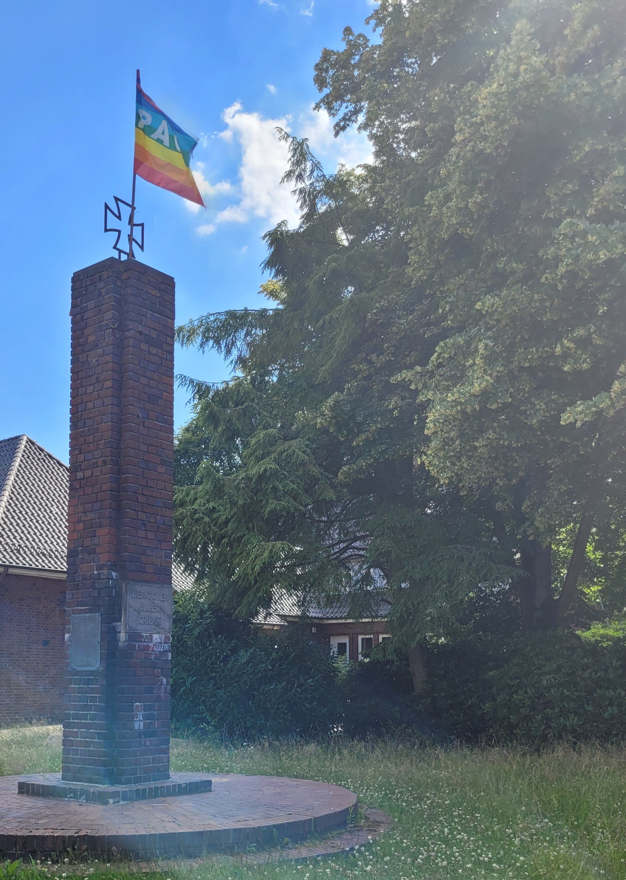 Auf einer kleinen, grünen, Gänseblümchenbewachsenen Wiese ist ein niedriger, gemauerter Kreis verlegt ,auf dem ein schmaler, eckiger Turm aus Klinker steht, ähnlich einem Schorstein. Oben auf dem Turm ist ein 'Eisernes Kreuz' angebracht, daran ist eine Ragenbogenfahne mit dem Schriftzug 'Pace' gehisst. Sie wwht im Wind. Im unteren Drittel des Turmes sind zwei Metalltafeln angebracht. Die eine ist unleserlich, auf der anderen steht:"Den Toten aller Kriege".
Im Hintergrund stehen große, sommergrüne Laubbäume und niedrige Häuser. Auf einer kleinen, grünen, Gänseblümchenbewachsenen Wiese ist ein niedriger, gemauerter Kreis verlegt ,auf dem ein schmaler, eckiger Turm aus Klinker steht, ähnlich einem Schorstein. Oben auf dem Turm ist ein 'Eisernes Kreuz' angebracht, daran ist eine Ragenbogenfahne mit dem Schriftzug 'Pace' gehisst. Sie wwht im Wind. Im unteren Drittel des Turmes sind zwei Metalltafeln angebracht. Die eine ist unleserlich, auf der anderen steht:"Den Toten aller Kriege".
Im Hintergrund stehen große, sommergrüne Laubbäume und niedrige Häuser.