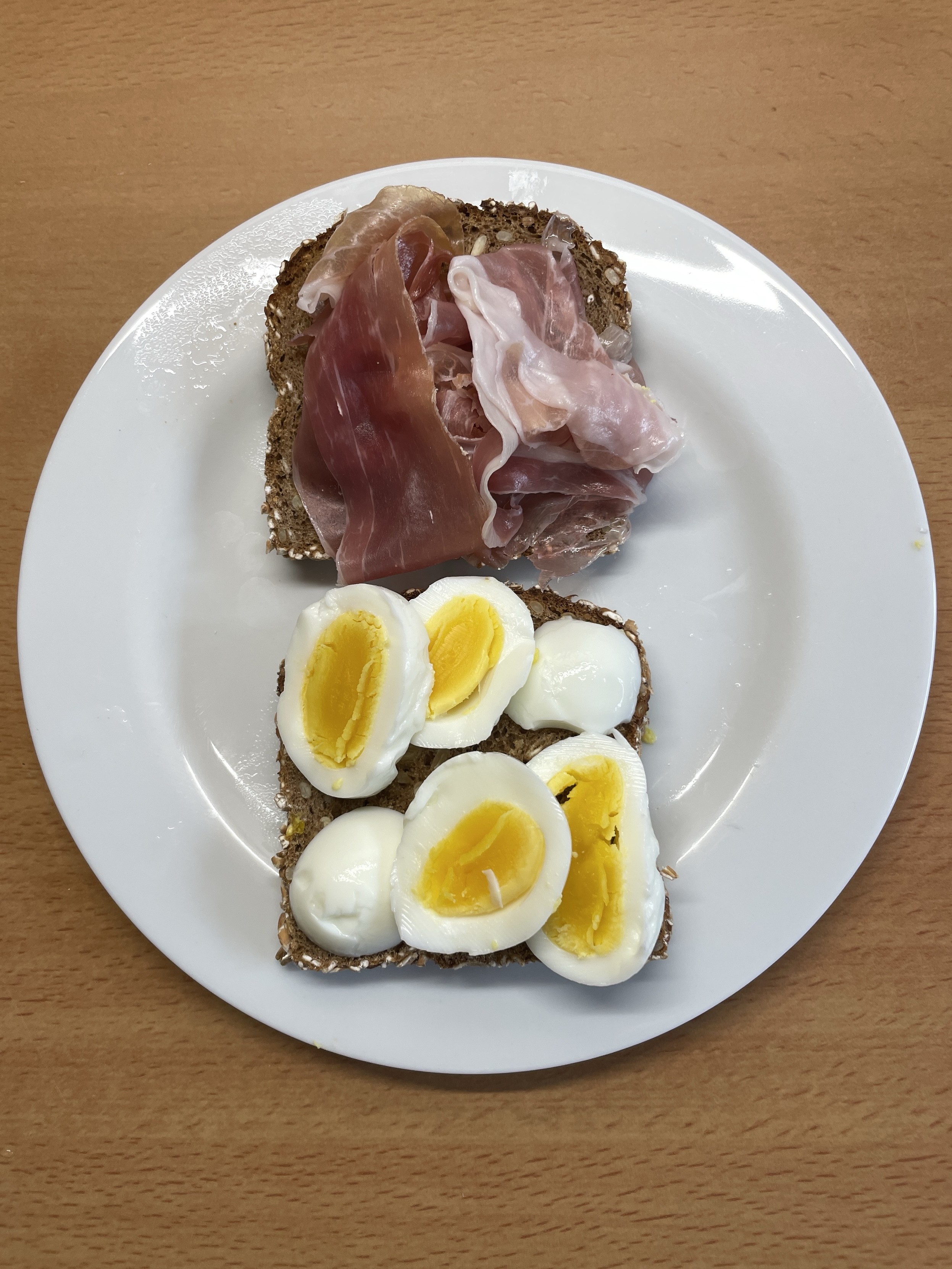 Ein belegtes Brot mit Schinken. Ein belegtes Brot mit Ei. Beide liegen auf einem weißen Teller. Im Hintergrund die Arbeitsfläche eine Küche.