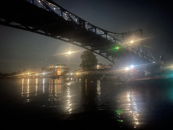 Das Bild zeigt eine nächtliche Szene im Hafen, auf dem ein Metallbrückenkranz über die Wasserfläche spannt. Die Brücke ist mit Lichtern ausgestattet, die auf das Wasser reflektieren und eine glitzernde Spiegelung erzeugen. Im Hintergrund ist ein Gebäude mit warmen Lichtern zu sehen, das an den Uferpromenade angebaut ist.  Die Umgebung ist in Dunkelheit gehüllt, mit einigen Lichtern, die durch die Nebelschwaden sichtbar sind. Die Atmosphäre ist ruhig und geheimnisvoll, mit dem Spiel von Licht und Schatten, das die Szene belebt.