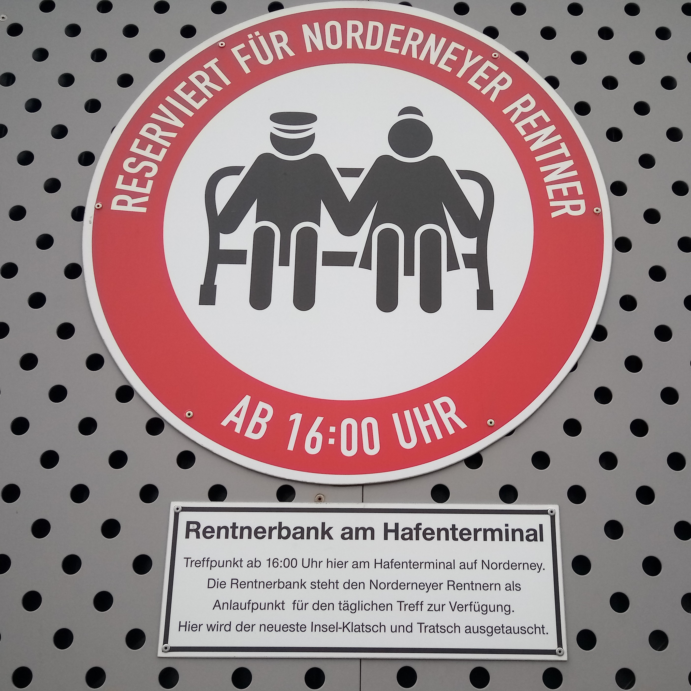 Quadratisches Format, ein rundes Schild, mittig eine Grafik mit zwei Personen auf einer Parkbank. Außen roter, breiter Rand, darin steht in weißer Schrift: RESERVIERT FÜR NORDERNEYER RENTNER AB 16:00 UHR.
Darunter weißes eckiges Schild mit der Erklärung: Rentnerbank am Hafenterminal