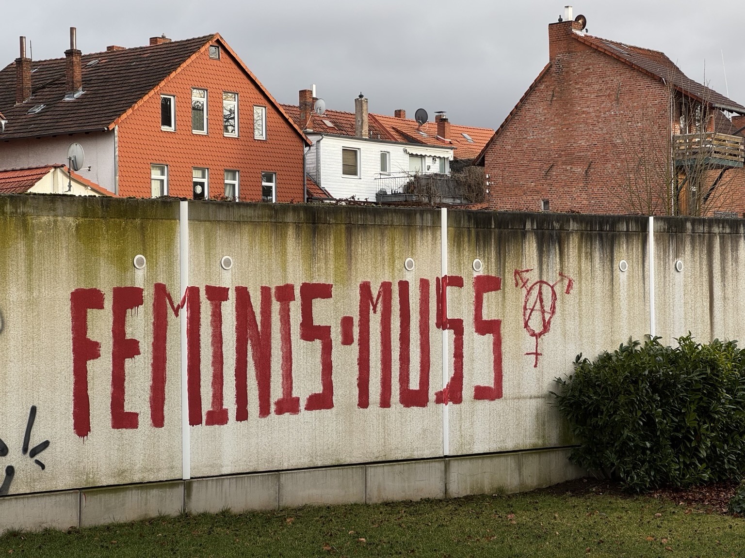 Eine Betonwand zeigt Graffiti mit der Aufschrift „FEMINIS*MUSS“ in fetten roten Buchstaben, begleitet von einem Symbol, das die weiblichen und Transgender-Symbole mit einem Anarchie-Zeichen kombiniert. Im Hintergrund befinden sich Wohngebäude mit markanten orangefarbenen Dächern.