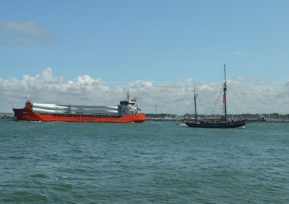 Ein Schiff mit rotem Rumpf, beladen mit Rotoren für Windkraftwerke, rechts in Gegenrichtung ein kleines Segelschiff, das unter Motor fährt. 