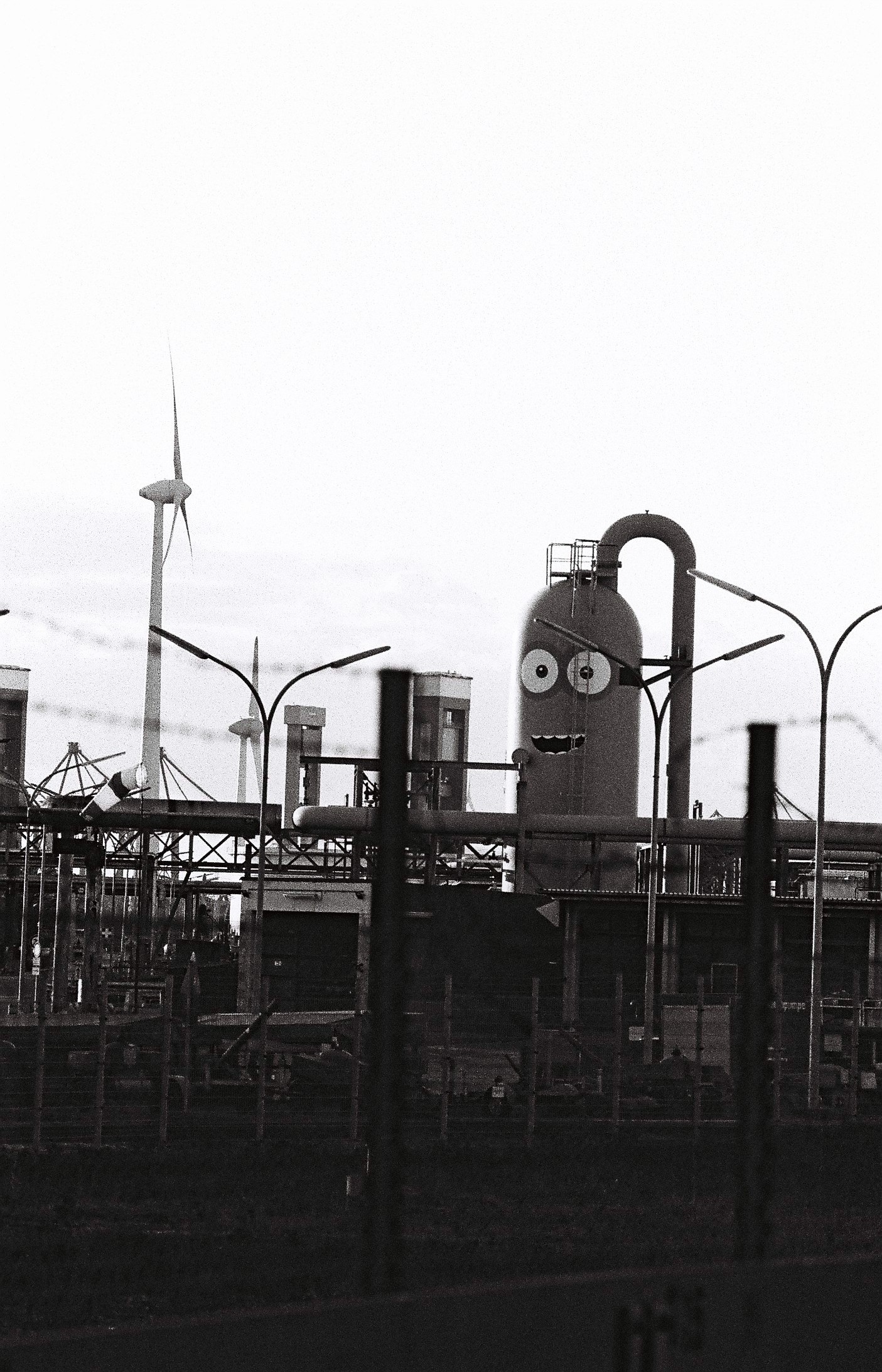 Eine Industrianlage mit einem Turm, der dank seiner Bemalung wie ein Minion aus „Ich, einfach unverbesserlich“ aussieht. Im Hintergrund sieht man einen Teil einer Hubbrücke und zwei Windräder.